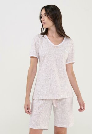 Mujer joven con camiseta de manga corta de color rosa claro con estampado y pantalones cortos a juego hasta la rodilla, mirando hacia su izquierda con un fondo liso.