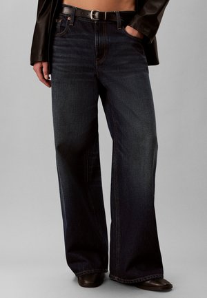 Person iført mørkeblå jeans med vide ben, sort bælte, sort læderjakke og sorte sko, den ene hånd i lommen og den anden ved siden.