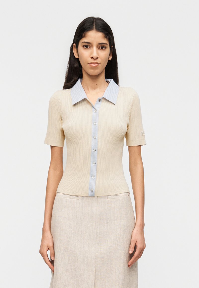 Γυναίκα με μακριά σκούρα μαλλιά, που φοράει ένα μπεζ ριγωτό μπλουζάκι με κοντά μανίκια και κουμπιά, με ανοιχτό μπλε γιακά και ταιριαστή μπεζ φούστα, σε λευκό φόντο.