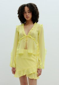 Geel transparant blouse met gerimpelde randen en een strikfront ontwerp, gecombineerd met een bijpassende gerimpelde rok. Lichtgewicht, vloeiend materiaal.