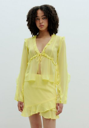 Geel transparant blouse met gerimpelde randen en een strikfront ontwerp, gecombineerd met een bijpassende gerimpelde rok. Lichtgewicht, vloeiend materiaal.