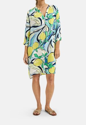 Femme portant une robe ample mi-longue avec un imprimé coloré de citrons et de feuilles, assortie à des sandales plates beiges sur fond blanc.