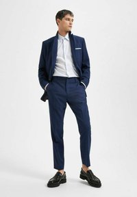 Tamsiai mėlynas kostiumas su struktūrizuotu švarku ir lieknomis kelnėmis. Tekstūruota medžiaga, balta marška ir juodi odiniai batai. Elegantiškas dizainas ir pritaikymas.
