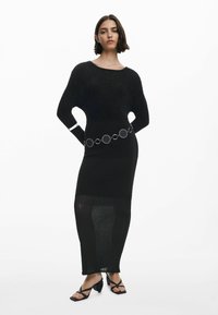Vestito nero a maglia con texture a coste, maniche lunghe e silhouette aderente, accentuato da una cintura metallica circolare. Indossato con sandali neri con cinturini.