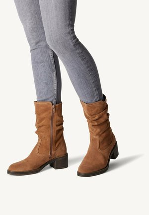 Bottines en suède marron avec un design ample, fermeture éclair, et un petit talon carré, portées avec un jean gris ajusté.