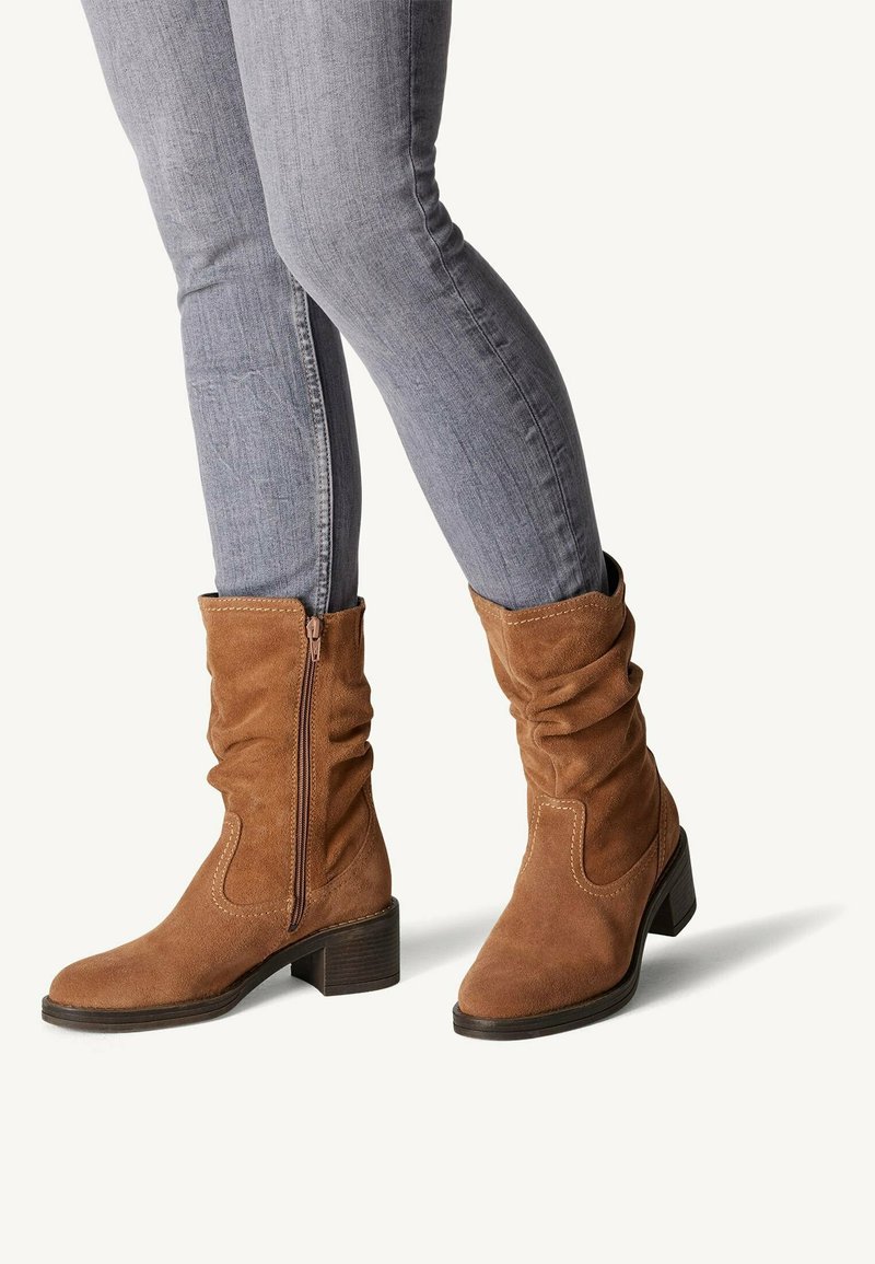 Brown suede ankle boots with a slouchy design, zip closure, and a low block heel, worn with gray fitted jeans.