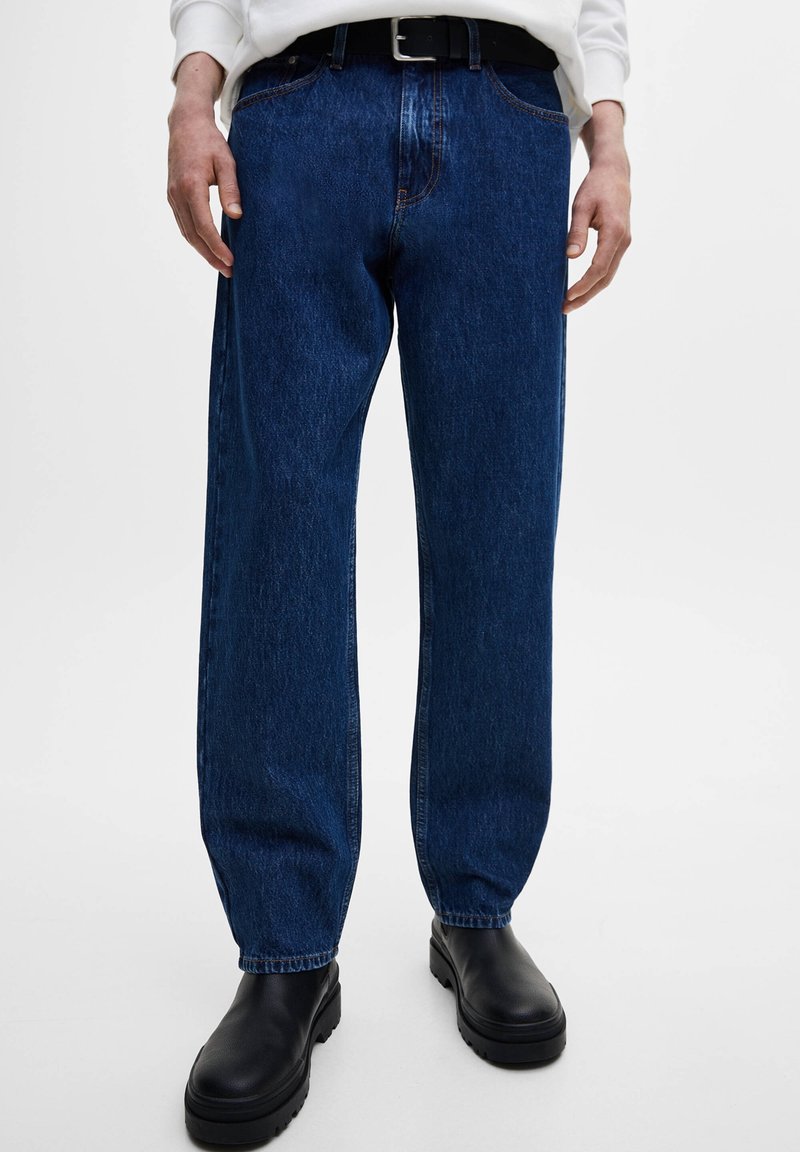 Vaqueros de pierna recta en azul oscuro con un acabado de denim texturizado, con costuras naranjas y un cinturón de cuero negro. Combinados con botas de plataforma negras.