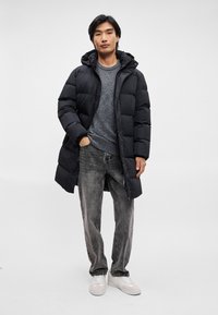 Schwarze Daunenjacke mit Kapuze, ausgestattet mit einem gesteppten Design. Grauer Pullover darunter, kombiniert mit grauen Jeans und weißen Turnschuhen.