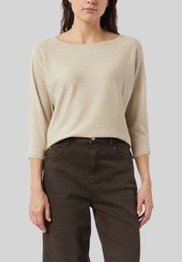 Beżowa, błyszcząca bluzka z długim rękawem o szerokim dekolcie, zestawiona z ciemnobrązowymi spodniami z wysokim stanem, charakteryzującymi się luźnym krojem i miękką fakturą.