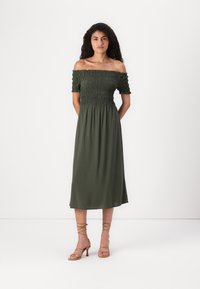 Olivgrön axelbandslös klänning med smockad bodice och svävande midiklänning, kombinerad med ljusbruna sandaler med klack och remmar.