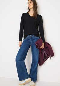 Musta pitkähihaisen yläosa, siniset leveälahkeiset farkut ja beigejä tennareita sinisillä yksityiskohdilla. Kädessä viininpunainen takki, minimalistinen muotoilu ja rento istuvuus.