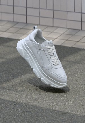 Białe sneakersy z grubą podeszwą i sznurowadłami, unoszące się nad asfaltem obok kafelkowanej ściany, prezentujące nowoczesny design i branding.