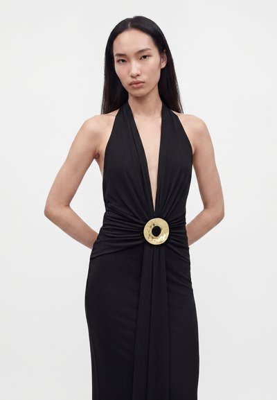 Woman with long black hair wearing a sleeveless black halter dress featuring a deep V-neck and a large round gold belt buckle at the waist.