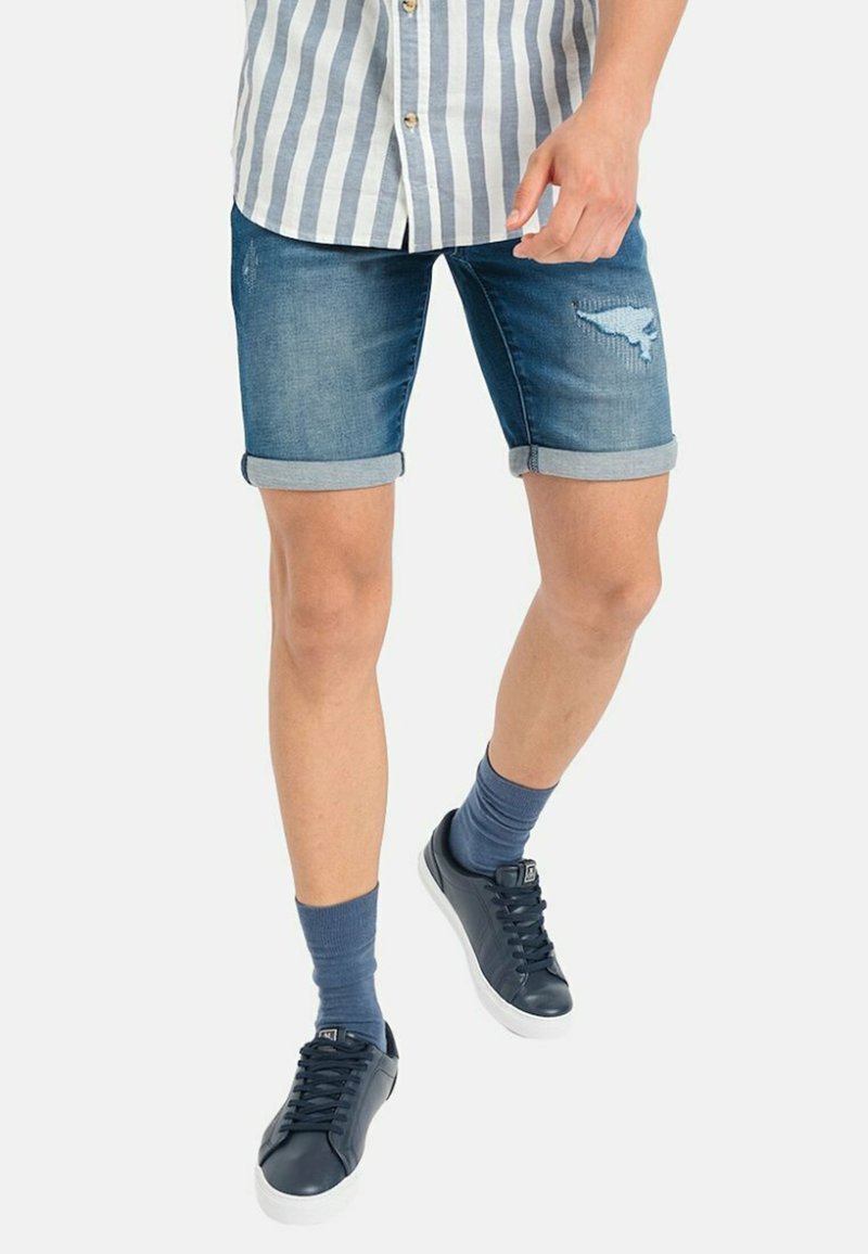 Shorts de mezclilla con un acabado desgastado, puños doblados, color azul claro. Combinados con zapatillas oscuras y calcetines hasta la altura del tobillo azul. Camiseta a rayas visible.