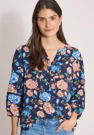 Mujer con el pelo largo y castaño lleva una blusa azul oscuro con flores rosas y azules, y vaqueros azules, de pie contra un fondo liso.