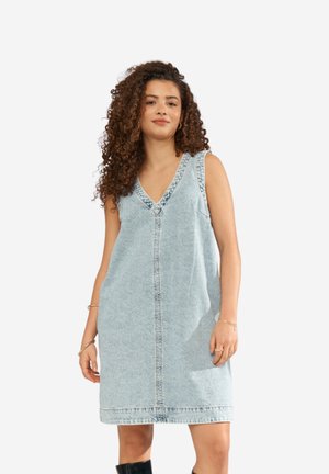 Žena s kučeravými vlasmi, oblečená v bezrukávových svetlomodrých rifľových šatách a čiernych čižmách, stojaca pred jednoduchým pozadím.