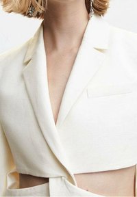 Femme portant un blazer court écru avec des revers crantés, présentant une découpe à l'avant et de longues boucles d'oreilles argentées pendantes.