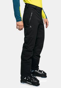 Schwarze Skihosen im Slim-Fit, mit Reißverschlusstaschen und elastischem Bund, getragen über schwarzen Skischuhen mit blauen Akzenten.