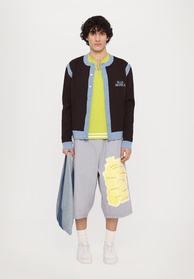 Black and light blue varsity jacket, neon green shirt, gray shorts with yellow graphics, white sneakers, holding a denim jacket.