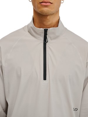 Man draagt een lichtgrijze pullover met kwartrits, zwarte ritssluiting en klein "LO" logo aan de rechteronderkant.