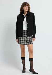 Black button-up shirt with embellished pockets paired with a checkered mini skirt. Model wears sheer black knee-high socks and black heels.