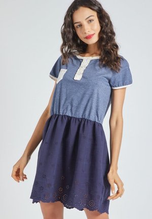 Mujer con cabello ondulado que lleva un vestido azul de manga corta con borde de encaje y falda bordada, posando frente a un fondo claro y sencillo.