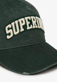 Gorra de béisbol de algodón verde con visera curva y bordado en relieve de "SUPERDRY" en color crema. La tela muestra una textura suave y costuras visibles.