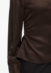 Brown satin blouse with long sleeves, featuring ruching on the side and a smooth texture. The design has a fitted silhouette and flared hem.