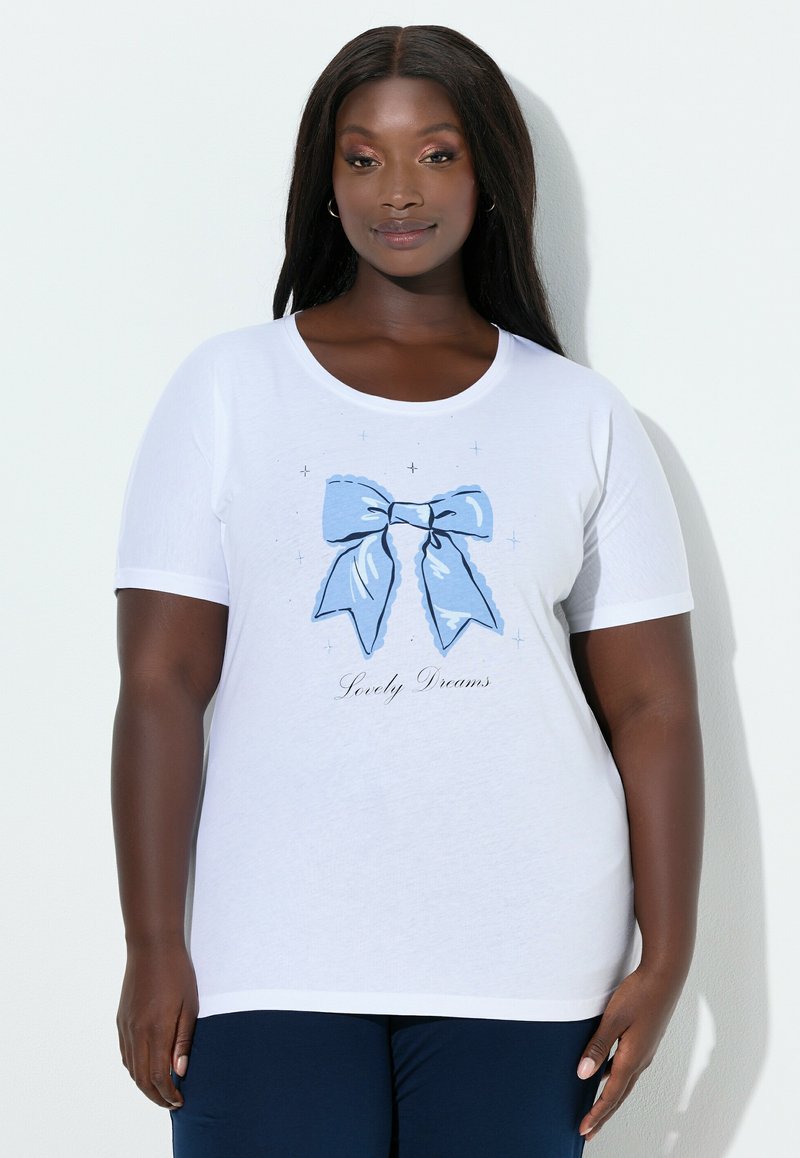 Femme grande taille portant un t-shirt blanc avec un grand nœud bleu et le texte « Lovely Dreams », assorti d'un pantalon bleu foncé, debout contre un mur blanc.