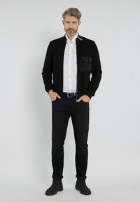 Czarna kurtka zapinana na zamek z kieszonką na klatce piersiowej, w połączeniu z białą koszulą zapinaną na guziki i czarnymi dżinsami o dopasowanym kroju. Szare buty dopełniają strój.