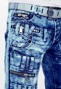 Jeans in denim di colore blu chiaro con multiple cerniere, una toppa trapuntata e diversi design delle tasche. Presentano una finitura testurizzata e dettagli cuciti.