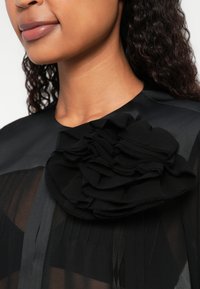 Blusa nera trasparente con una grande accentatura di fiore testurizzato sulla spalla, con tessuti sovrapposti che creano profondità e contrasto nel design.