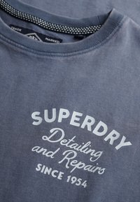 Camiseta de algodón gris con escote redondo. En la parte delantera presenta texto blanco: "SUPERDRY Detalles y Reparaciones Desde 1954." Textura suave.