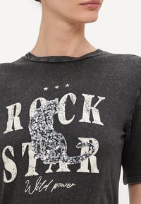 Pilkas marškinėliai su nusidėvėjusia "ROCK STAR" spauda, papuošti sidabrinėmis blizgučių katės detalėmis ir balta tekstu "Wild power."