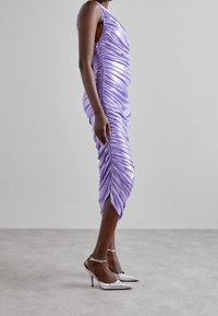 Levandulové saténové šaty s vertikálním řasením, bez rukávů a rozparkem na spodním lemu. V kombinaci se stříbrnými lodičkami se špičatou špičkou a řemínky kolem kotníku.