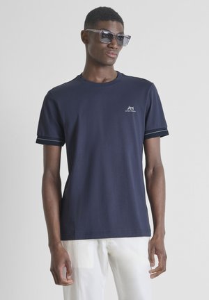 Uomo che indossa una t-shirt con logo a maniche corte blu scuro, pantaloni bianchi e grandi occhiali da sole quadrati, in piedi contro uno sfondo chiaro e uniforme.