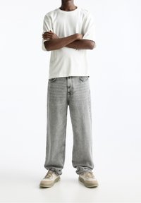 Une personne debout, les bras croisés, portant un t-shirt blanc uni, un jean gris ample et des baskets beiges, sur un fond blanc.