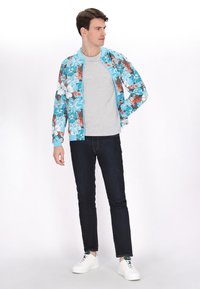 Giubbotto bomber leggero con stampa floreale in blu, grigio e rosso, abbinato a una t-shirt grigia, jeans scuri e sneakers bianche.