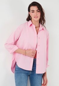 Light pink button-up shirt with long sleeves, loose fit, and side slits. Paired with high-waisted denim jeans. Simple collar and soft texture.