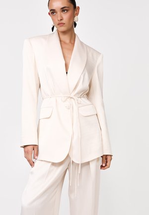 Nő, aki szabott törtfehér öltönyt visel öves blézerrel és hozzá illő, bő szárú nadrággal, egyszerű világos háttér előtt.