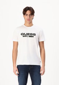 Tricou alb din bumbac cu imprimare neagră "GUESS EST.1981" pe față, mâneci scurte și guler rotund. Asortat cu blugi bleu închis.