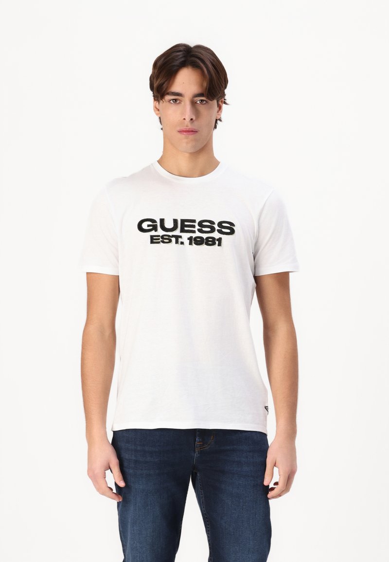 Tricou alb din bumbac cu imprimare neagră "GUESS EST.1981" pe față, mâneci scurte și guler rotund. Asortat cu blugi bleu închis.