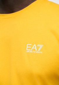 Κοντινό πλάνο κίτρινης μπλούζας με λευκό λογότυπο EA7 Emporio Armani στο στήθος, εμφανίζοντας μέρος του λαιμού και του ώμου ενός ατόμου.
