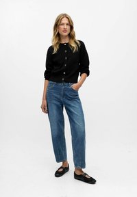 Cardigan nero a maglia con scollatura rotonda e chiusura con bottoni, abbinato a jeans in denim blu dalla vestibilità comoda. Modella che indossa ballerine nere, in piedi in modo casual.