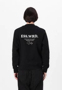 Black crewneck sweatshirt featuring white text: "ESS.WRD.," "SINCE NINETEEN NINETY," and "ESSENTIALS DEPT. Studio" on the back.