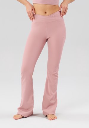 Femme portant des leggings évasés roses à taille haute et côtelés, les mains reposant sur les hanches, debout pieds nus sur un fond clair.