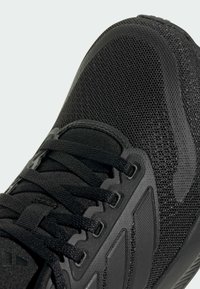 Černá sportovní bota s texturovaným síťovinovým svrškem, plochými černými tkaničkami a gumovou podrážkou s geometrickým vzorem pro lepší trakci.