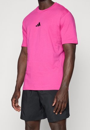 Homme portant un t-shirt rose vif à manches courtes avec un petit logo noir sur la poitrine et un short de sport noir sur fond blanc.
