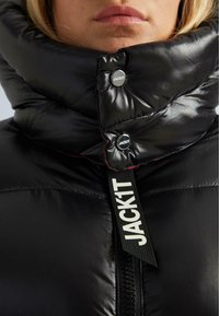 Primo piano di una persona che indossa un giubbino in nylon lucido nero con colletto alto, zip e bottoni etichettati "JACKIT".
