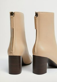 Botas de tobillo de cuero beige con punta redonda, una cremallera lateral visible y un tacón cuadrado marrón. Acabado liso con un brillo sutil.